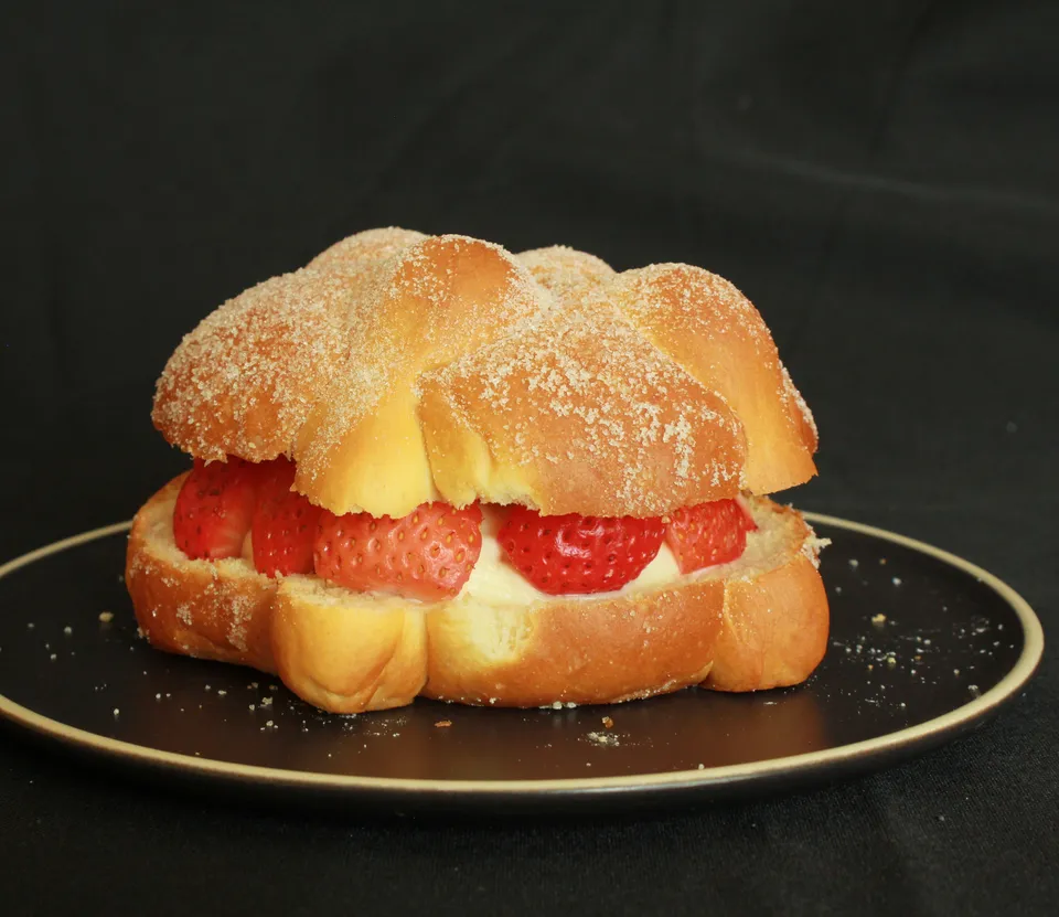 Pan de muerto abierto y relleno de fresas rebanadas y crema, en un plato negro