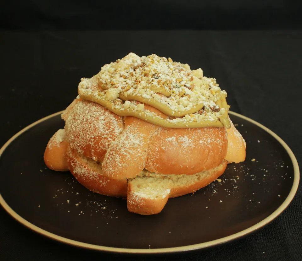 Pan de muerto relleno, con crema verde clara y pistache picado encima, en un plato negro